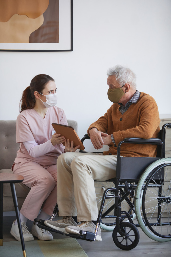Vertical full length portrait of young female nurse assisting senior man in wheelchair using digital tablet at retirement home, both wearing masks