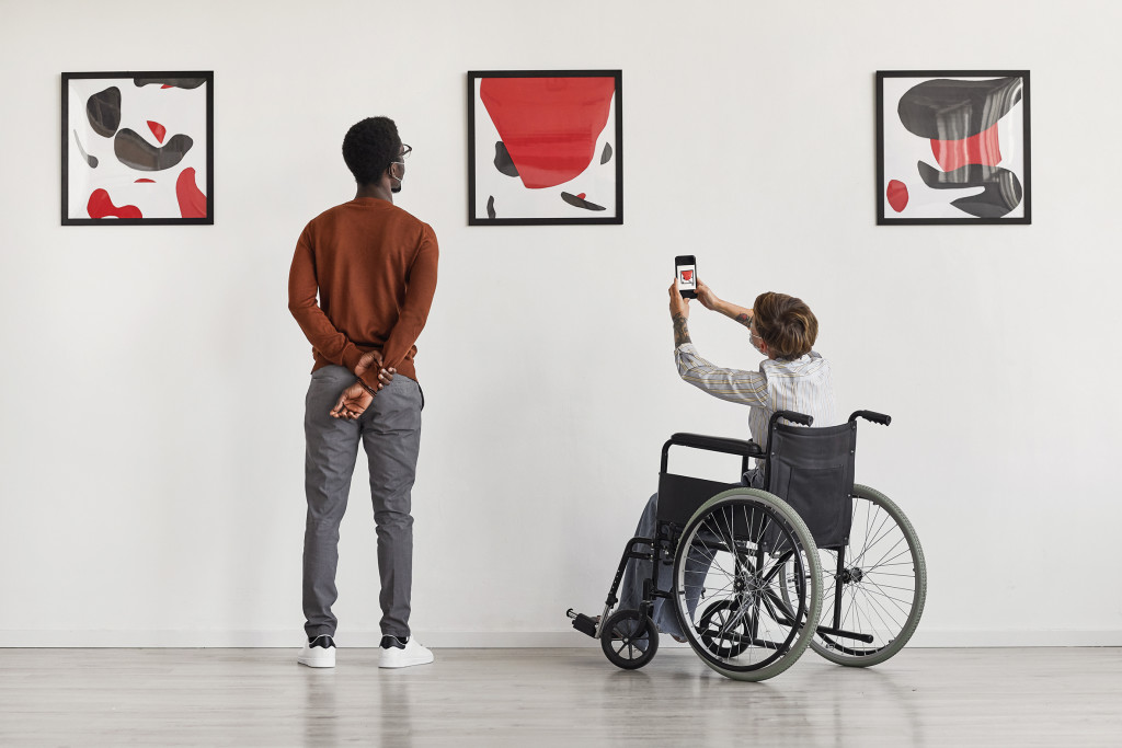 Back view portrait of young woman using wheelchair taking photo of artwork while visiting accessible museum, copy space