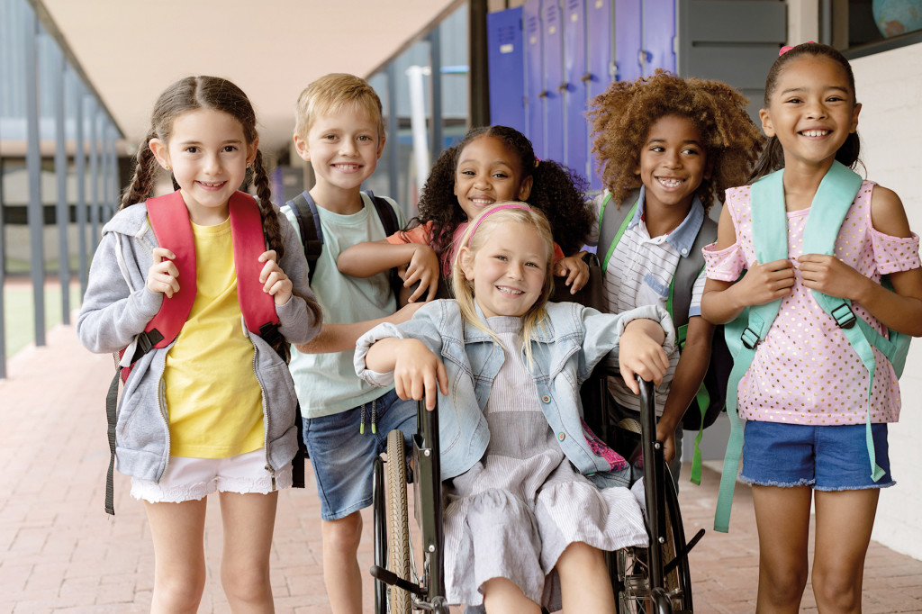 Front view of happy diverse school kids standing in  outside corridor at school while a Caucasian schoolgirl is sitting on wheelchair in foreground