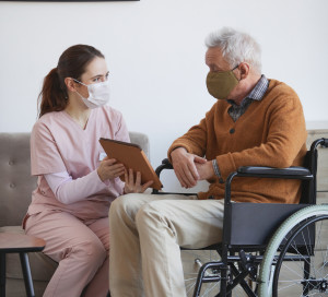 Vertical full length portrait of young female nurse assisting senior man in wheelchair using digital tablet at retirement home, both wearing masks