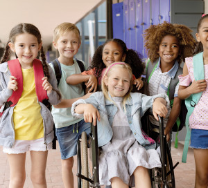 Front view of happy diverse school kids standing in  outside corridor at school while a Caucasian schoolgirl is sitting on wheelchair in foreground