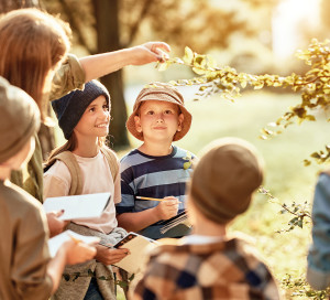 Group of curious school children with notebooks listening to their young female teacher while learning about nature together, looking at green leaf during ecology lesson in autumn forest on sunny day