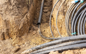 Thick walled plastic pipe and coiled armored cables in dug trench for underground power line installation under roadway. Soil excavation and cable laying preparation, selective focus