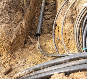 Thick walled plastic pipe and coiled armored cables in dug trench for underground power line installation under roadway. Soil excavation and cable laying preparation, selective focus